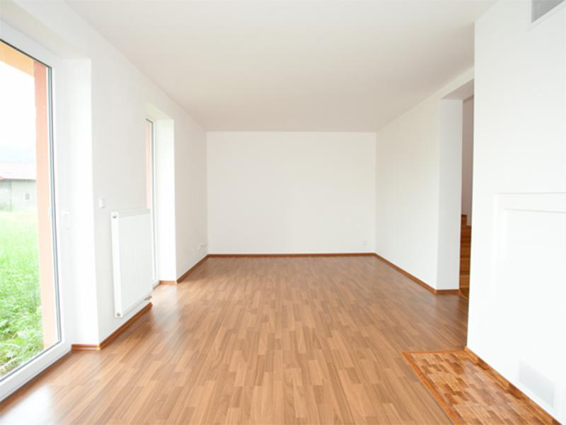 Puste, jasne wnętrze z panelami podłogowymi imitującymi drewno, białymi ścianami, grzejnikiem i oknem z widokiem na zieleń, ukazujące stan surowy po malowaniu.