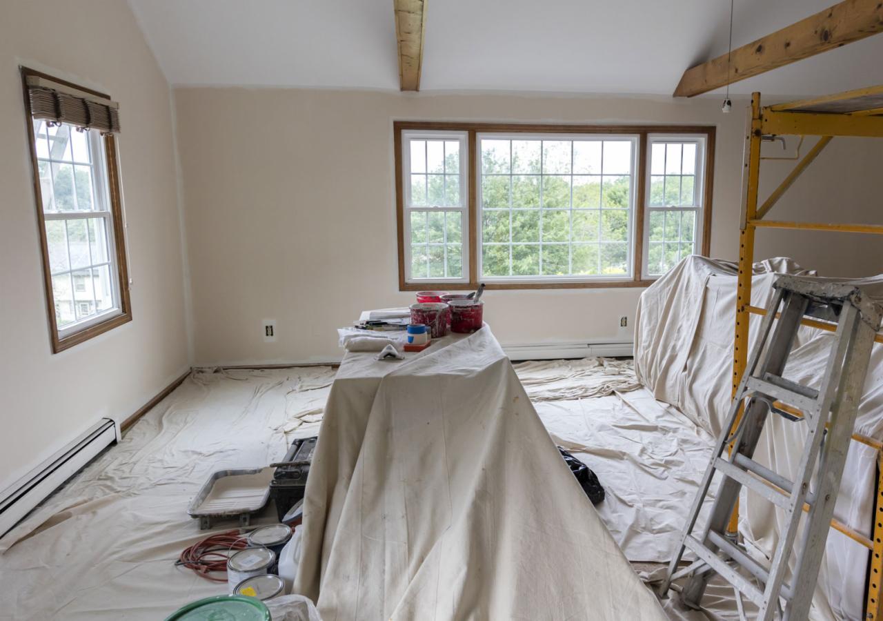 Pokój w trakcie malowania: ściany pokryte świeżą warstwą farby w odcieniu beżu, podłoga i meble zabezpieczone folią malarską, widoczne pędzle, wałki i puszki z farbą oraz drabina i rusztowanie.