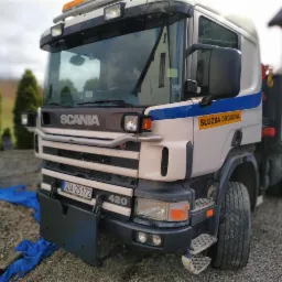 Ciężarówka Scania z napisem 'Służba Drogowa' zaparkowana na żwirowym podjeździe, z widocznym numerem rejestracyjnym i fragmentem niebieskiej plandeki.