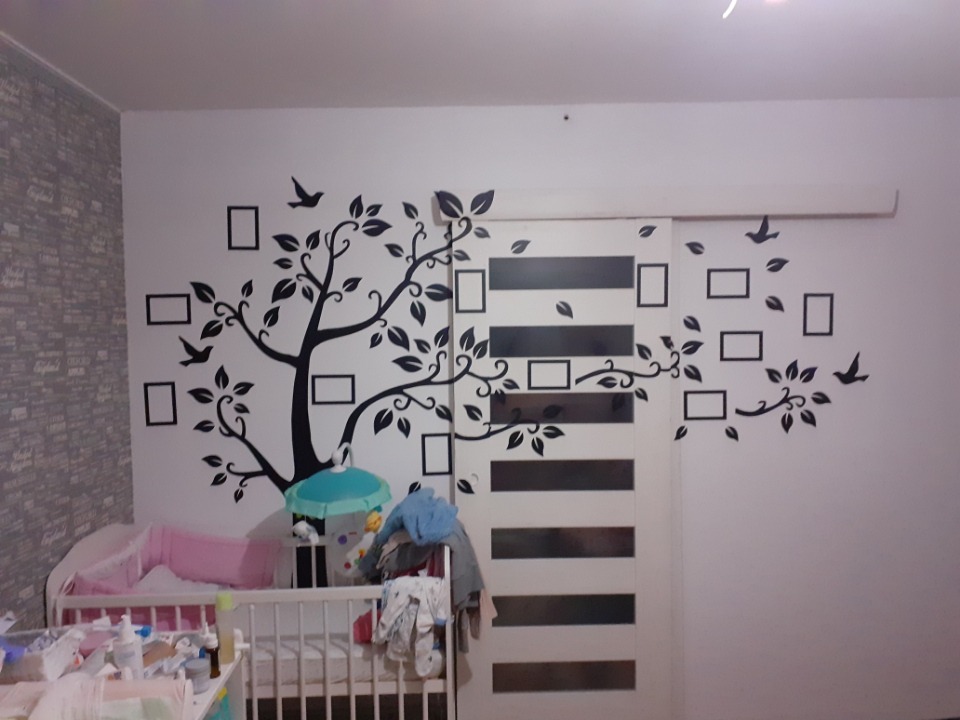 Czarny, dekoracyjny motyw drzewa z ramkami i ptakami na białej ścianie w pokoju dziecięcym, częściowo zasłaniający przesuwne drzwi szafy.