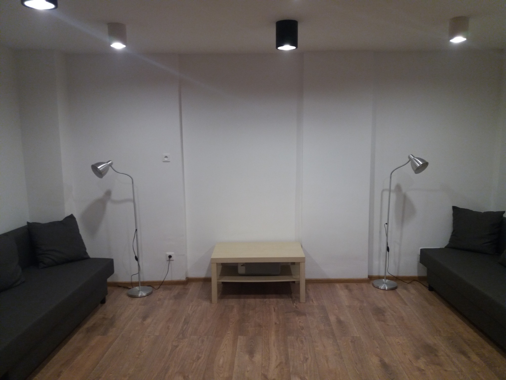 Pomalowane na biało ściany w pustym pokoju z drewnianą podłogą, dwiema szarymi sofami po bokach, niskim stolikiem kawowym na środku i lampami podłogowymi, gotowe do umeblowania.