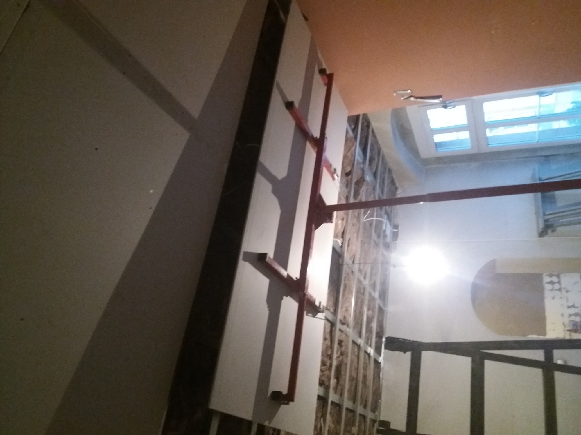 Montaż płyty gipsowo-kartonowej na ścianie z widoczną konstrukcją metalową i izolacją, podtrzymywanej przez czerwony stempel budowlany.