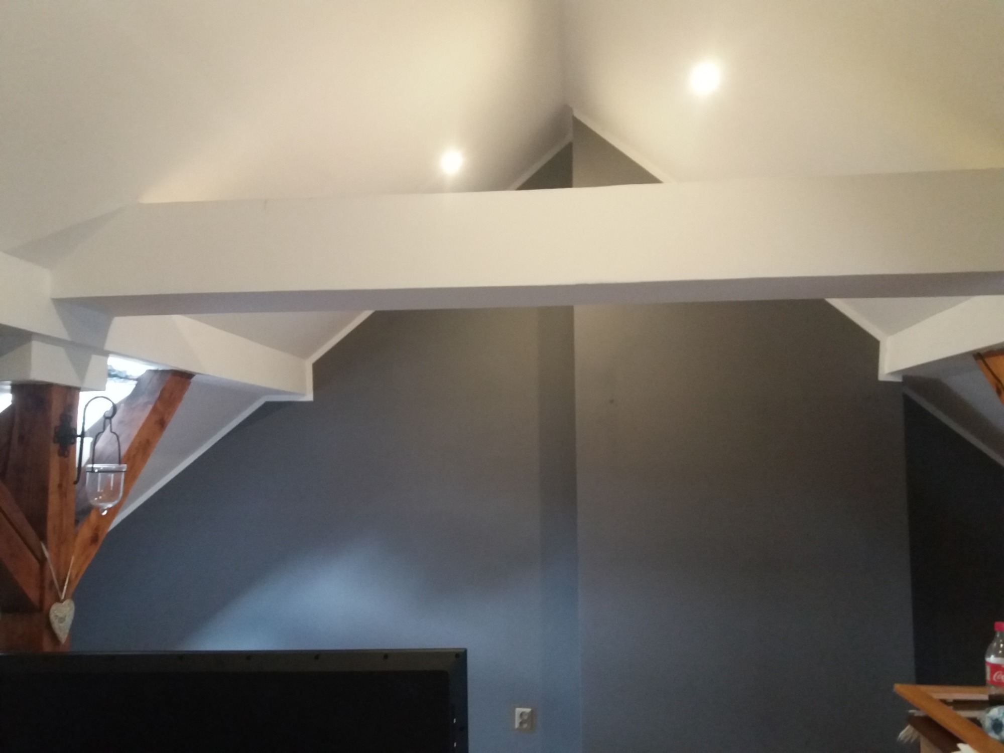 Szara ściana w pokoju na poddaszu po remoncie, widoczne białe belki stropowe i oświetlenie punktowe w suficie.