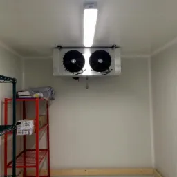 Wnętrze komory chłodniczej z zamontowanym agregatem chłodniczym pod sufitem, oświetlonym lampą jarzeniową, z czerwonym regałem z półkami po lewej stronie.