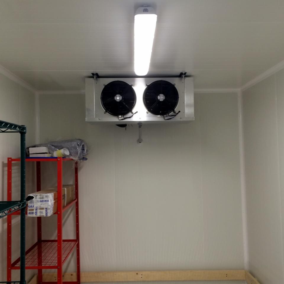 Wnętrze komory chłodniczej z zamontowanym agregatem chłodniczym pod sufitem, oświetlonym lampą jarzeniową, z czerwonym regałem z półkami po lewej stronie.