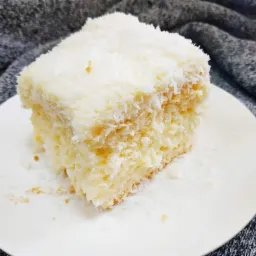 Kwadratowy kawałek ciasta kokosowego na białym talerzu, z widoczną strukturą warstw i posypką z wiórków kokosowych, na tle szarej dzianiny.