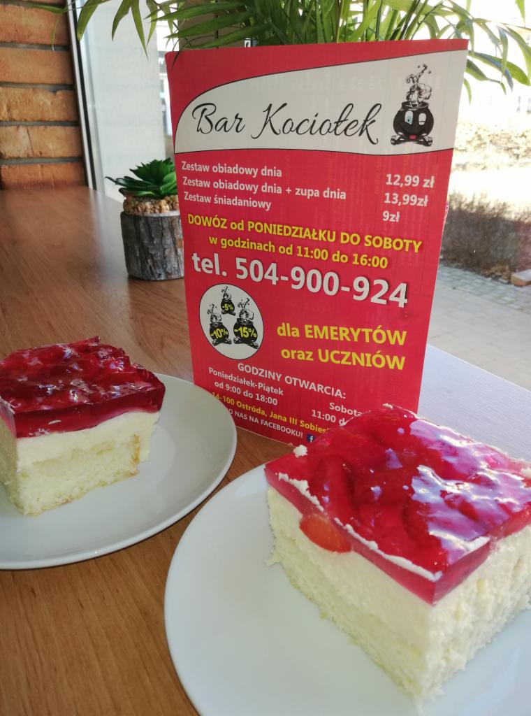 Dwa kawałki ciasta z galaretką truskawkową na białych talerzach, w tle menu baru 'Kociołek' z ofertą zestawów obiadowych i informacją o dowozie.