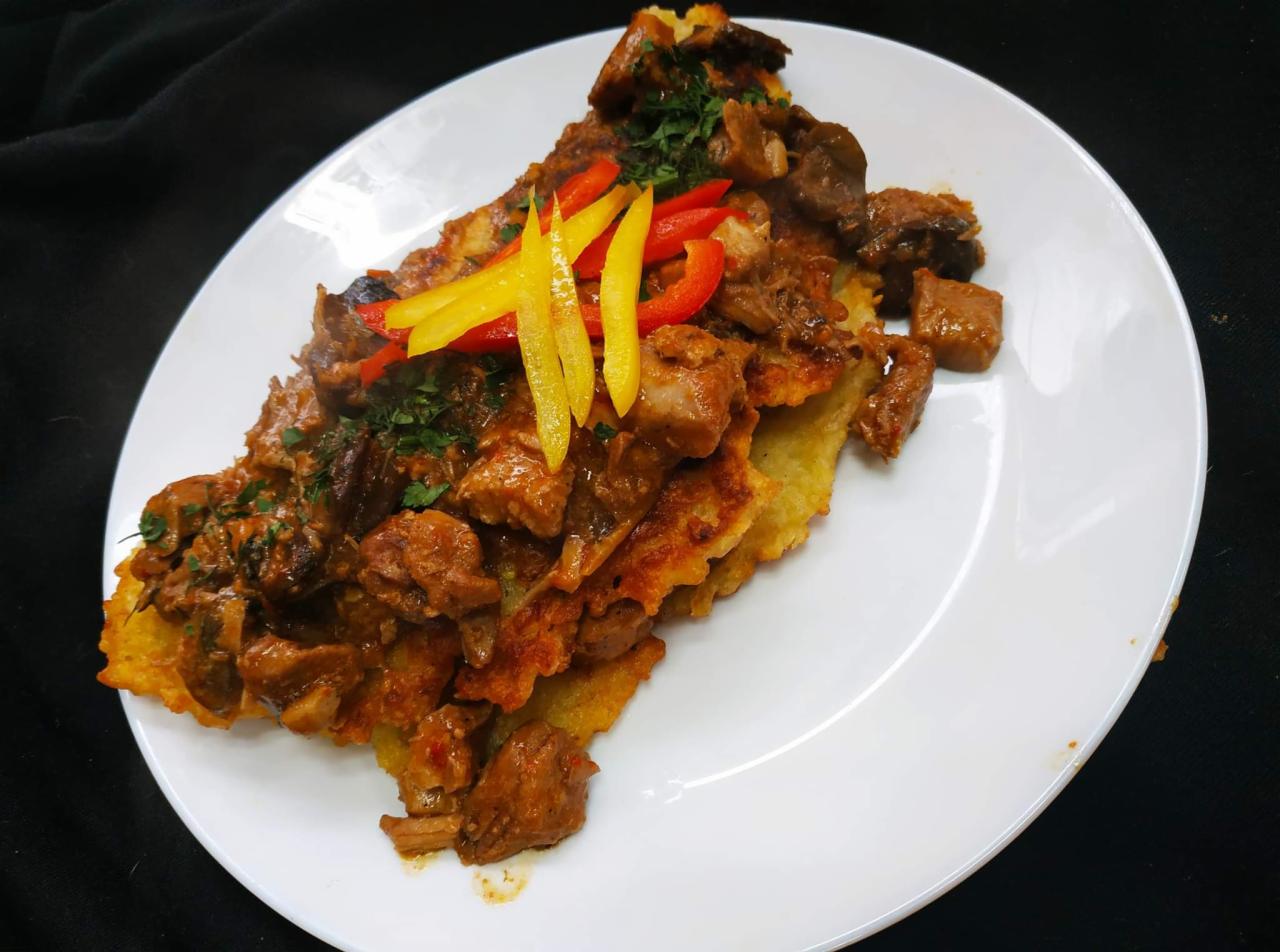 Placek ziemniaczany z gulaszem wieprzowym, udekorowany paskami papryki i natką pietruszki, podany na białym talerzu na czarnym tle.