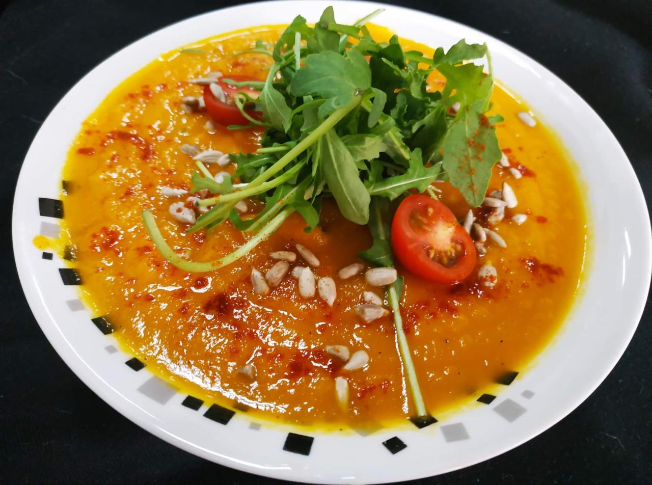Kremowa pomarańczowa zupa w białym talerzu z geometrycznym wzorem, udekorowana rukolą, połówkami pomidorków cherry i posypana ziarnami słonecznika oraz przyprawą.