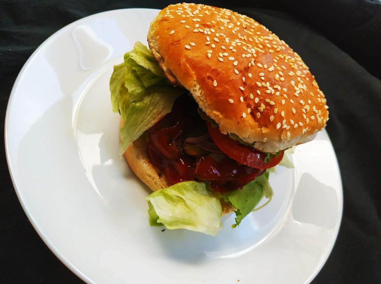 Apetyczny hamburger z sezamową bułką, świeżą sałatą, pomidorem i sosem na białym talerzu, sfotografowany z bliska na ciemnym tle.