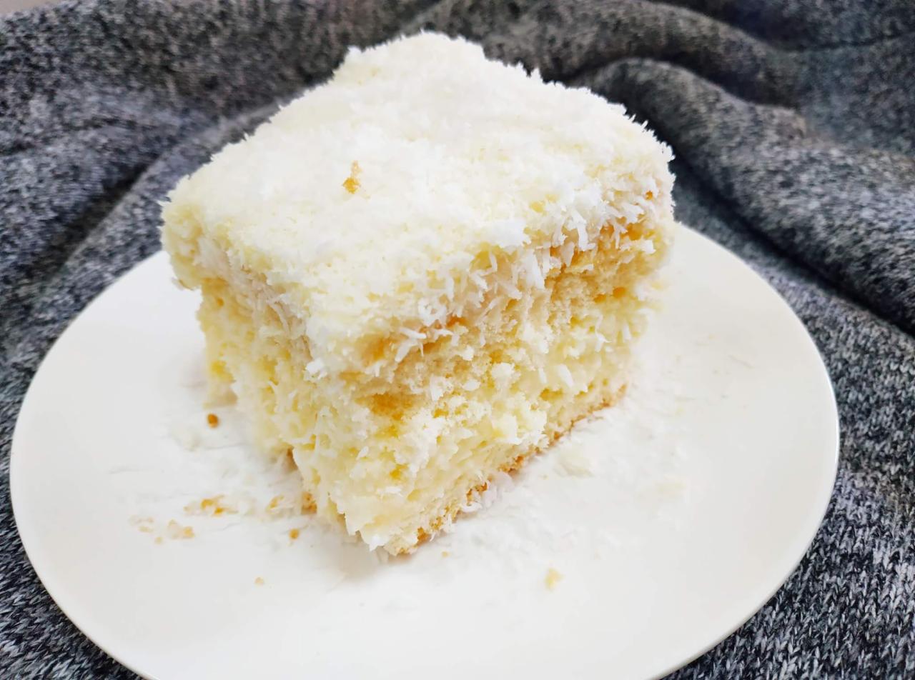 Kwadratowy kawałek ciasta kokosowego na białym talerzu, z widoczną strukturą warstw i posypką z wiórków kokosowych, na tle szarej dzianiny.