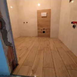 Łazienka w trakcie wykańczania: ułożona podłoga z paneli imitujących drewno, częściowo wykończone ściany z widocznymi instalacjami, surowy fragment ściany z odsłoniętą cegłą.