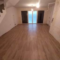 Salon w trakcie remontu z nowo ułożoną podłogą z paneli imitujących drewno, widoczne niedokończone ściany, okno balkonowe zabezpieczone folią i schody w stanie surowym.