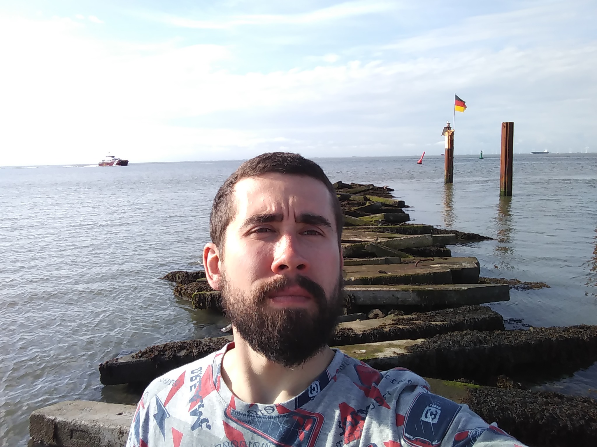 Mężczyzna robiący selfie na betonowym molo w morzu, w tle widoczny statek i niemiecka flaga na palu.