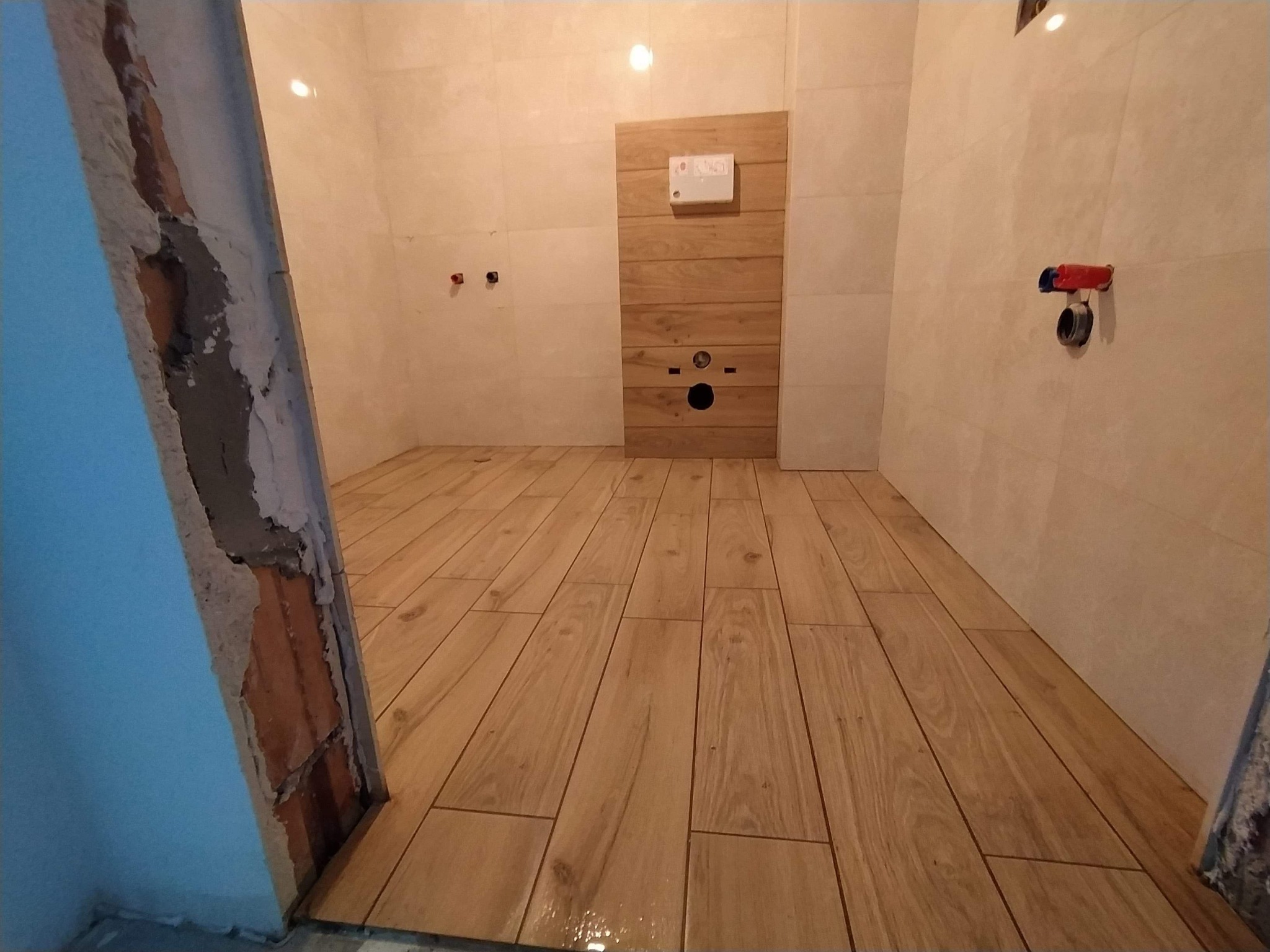 Łazienka w trakcie wykańczania: ułożona podłoga z paneli imitujących drewno, częściowo wykończone ściany z widocznymi instalacjami, surowy fragment ściany z odsłoniętą cegłą.