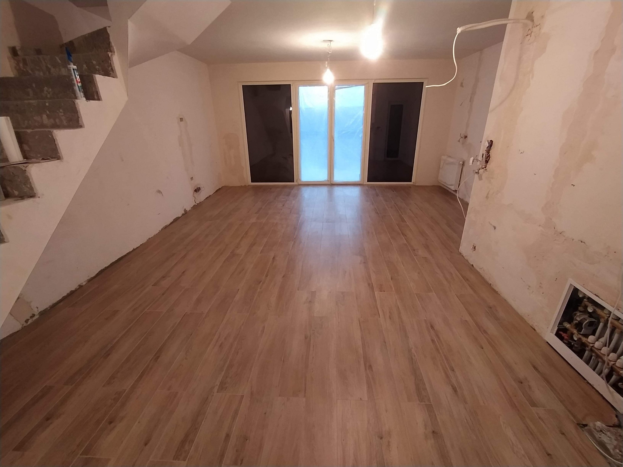 Salon w trakcie remontu z nowo ułożoną podłogą z paneli imitujących drewno, widoczne niedokończone ściany, okno balkonowe zabezpieczone folią i schody w stanie surowym.