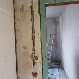 MatBud - Ściana w trakcie prac remontowych, widoczne fragmenty starego tynku, przewody elektryczne w bruzdach, częściowo zamontowana płyta gipsowo-kartonowa oraz metalowa drabina w tle.