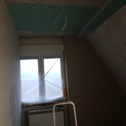 MatBud - Pokój na poddaszu w trakcie prac wykończeniowych: ściany po gładzi, okno zabezpieczone folią, częściowo zamontowana płyta karton gips na suficie, widoczny fragment drabiny.