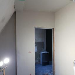 MatBud - Pokój po nałożeniu gładzi szpachlowej, widoczne niedoskonałości przed malowaniem, lampa halogenowa oświetla ścianę, w tle otwór drzwiowy do pomieszczenia przed remontem.