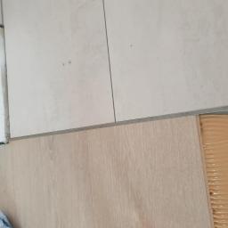 MatBud - Fragment podłogi z jasnych paneli, widoczny klej montażowy, łączenie z płytkami ceramicznymi i listwą progową.