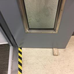 MatBud - Szare, pomalowane drzwi z metalową ramką wokół okienka, podparte drewnianym klinem na jasnej podłodze z żółto-czarną taśmą ostrzegawczą na framudze.