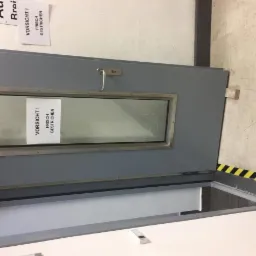 Otwarta szara metalowa brama z szybą i ostrzeżeniem 'Vorsicht! Frisch Gestrichen' (Uwaga! Świeżo malowane), widoczne czarno-żółte pasy ostrzegawcze na podłodze.