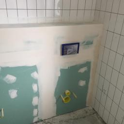 MatBud - Fragment ściany w łazience w trakcie prac wykończeniowych: zielona płyta gipsowo-kartonowa, częściowo pokryta białą masą szpachlową, widoczne żółte elementy instalacji hydraulicznej i białe płytki...