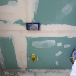 MatBud - Ściana z płyty karton gips, częściowo wyszpachlowana, z widocznymi puszkami instalacyjnymi Sigma i żółtymi rurami odpływowymi, w trakcie prac wykończeniowych.