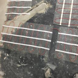 MatBud - Montaż mat grzewczych pod wylewkę na nierównym, szarym podłożu, widoczne przewody i fragment buta roboczego w rogu.