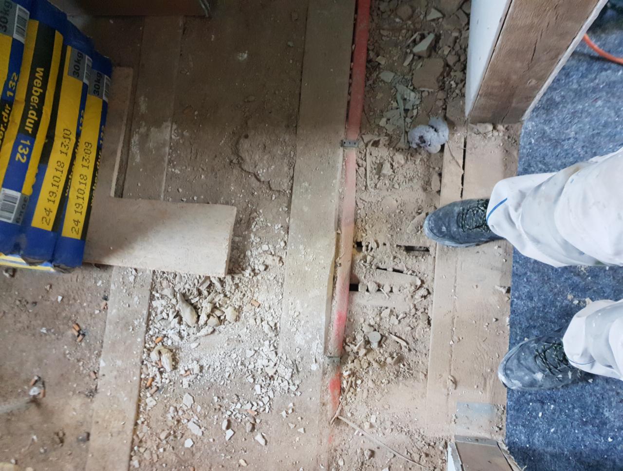 Przygotowanie podłoża pod nową podłogę: widok z góry na starą, drewnianą podłogę z resztkami gruzu, fragmentem czerwonej rurki, workami z zaprawą Weber.dur 132 i nogami osoby w kombinezonie roboczym.