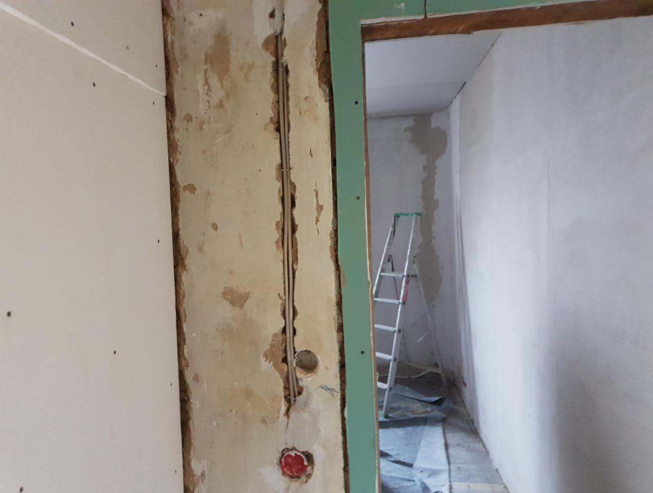 Ściana w trakcie prac remontowych, widoczne fragmenty starego tynku, przewody elektryczne w bruzdach, częściowo zamontowana płyta gipsowo-kartonowa oraz metalowa drabina w tle.