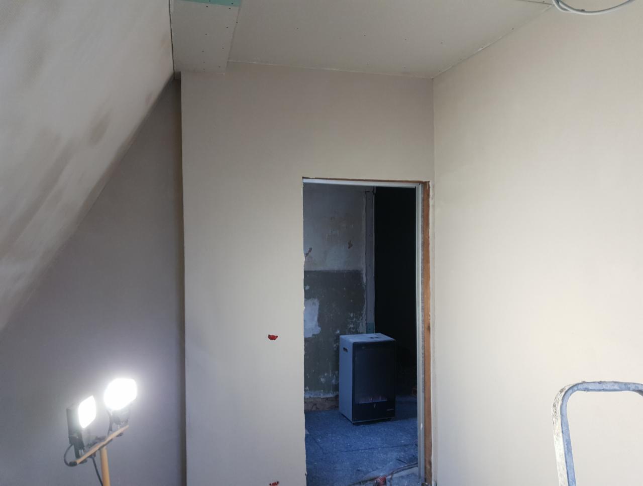 Pokój po nałożeniu gładzi szpachlowej, widoczne niedoskonałości przed malowaniem, lampa halogenowa oświetla ścianę, w tle otwór drzwiowy do pomieszczenia przed remontem.