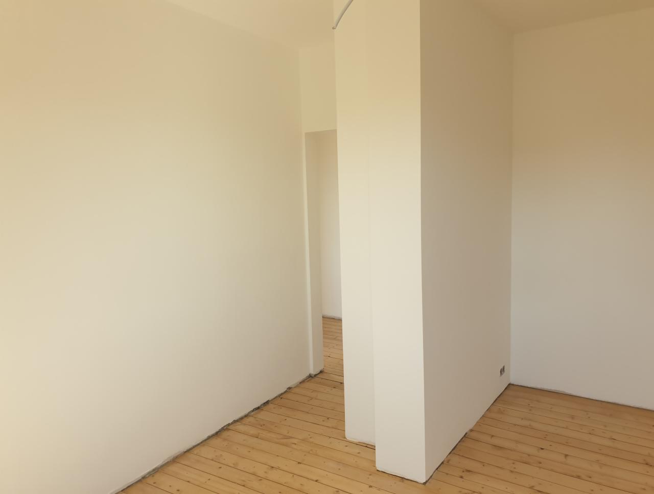 Jasne, świeżo pomalowane ściany w odcieniu kremowym w pustym pokoju z drewnianą podłogą, widoczne gniazdko elektryczne w dolnej części ściany.