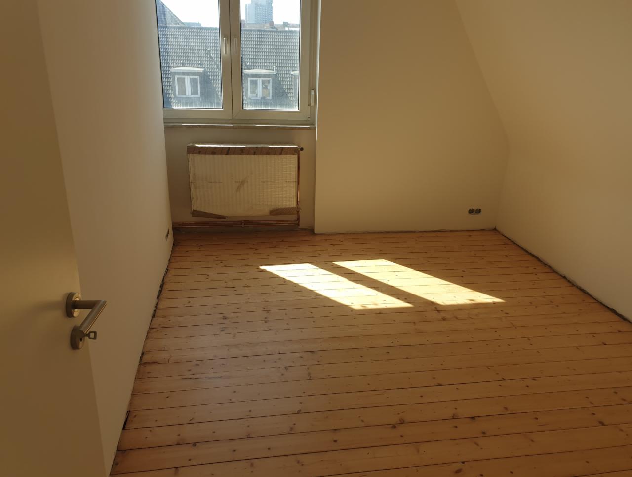 Jasne, puste pomieszczenie na poddaszu z drewnianą podłogą w jodełkę, świeżo pomalowanymi ścianami na kolor kremowy i widokiem na budynki przez okno.