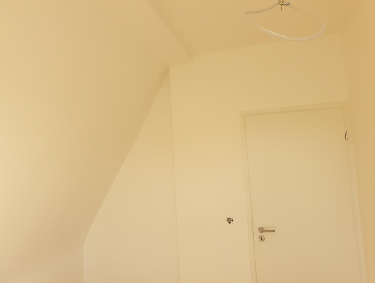 Jasne wnętrze z białymi ścianami i drzwiami, widoczny fragment skośnego sufitu i lampa wisząca w kształcie półkola.