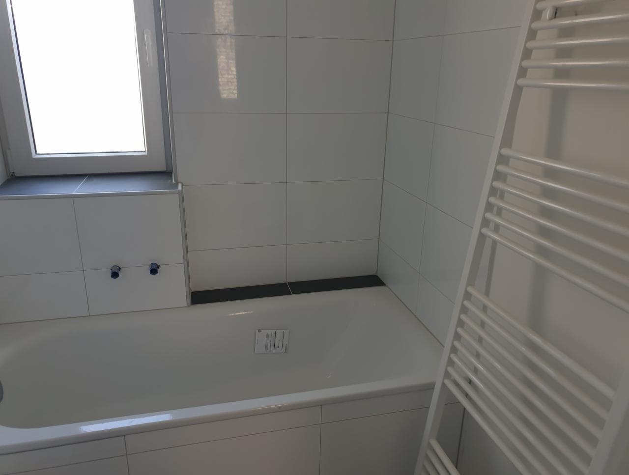 Biała wanna z zabudową pod oknem i grzejnikiem drabinkowym na tle białych płytek ściennych.