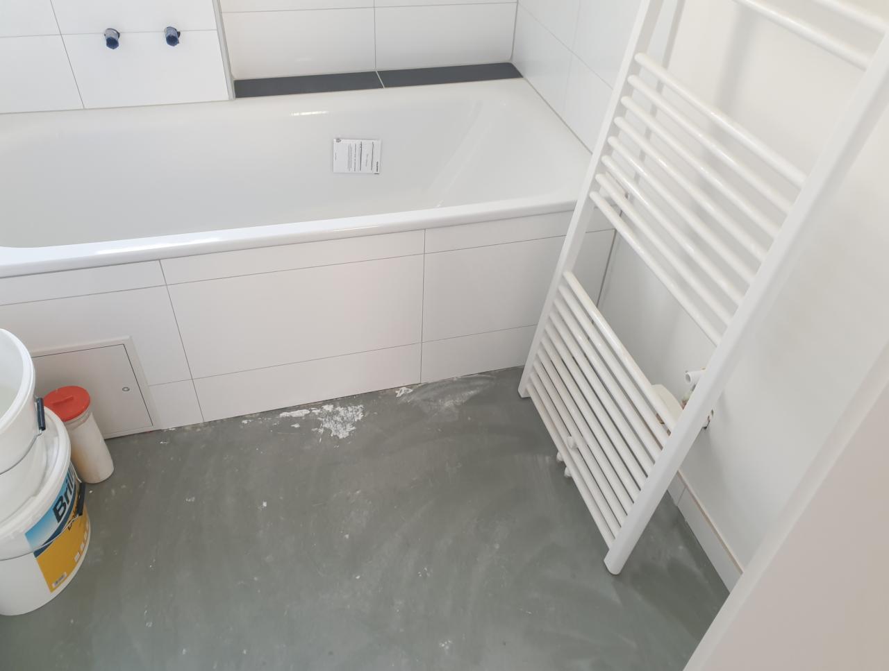 Biała wanna z białymi płytkami, grzejnik drabinkowy i wiadro farby na szarym, częściowo zabrudzonym podłożu, wskazujące na prace wykończeniowe w łazience.