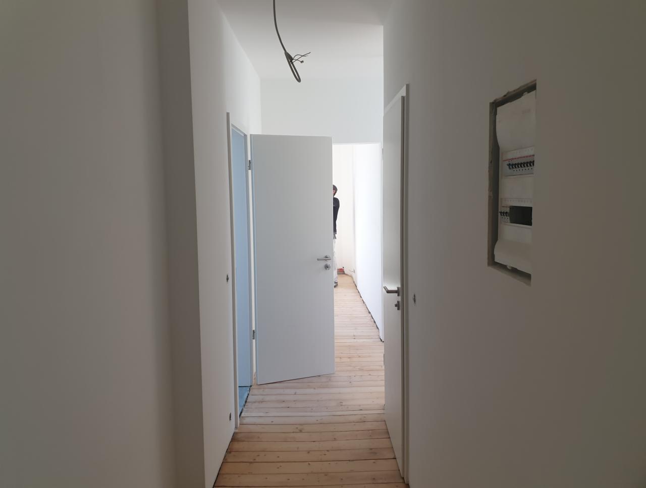 Białe, świeżo pomalowane wnętrze mieszkania w trakcie wykańczania, widoczne otwarte drzwi, drewniana podłoga i skrzynka elektryczna w ścianie.