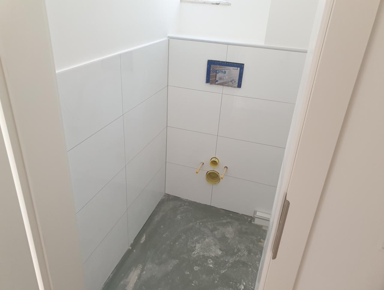 Wnętrze łazienki w trakcie remontu: białe, prostokątne płytki ułożone na ścianach, widoczne przyłącza kanalizacyjne i stelaż podtynkowy, surowa, betonowa podłoga, otwarte drzwi.