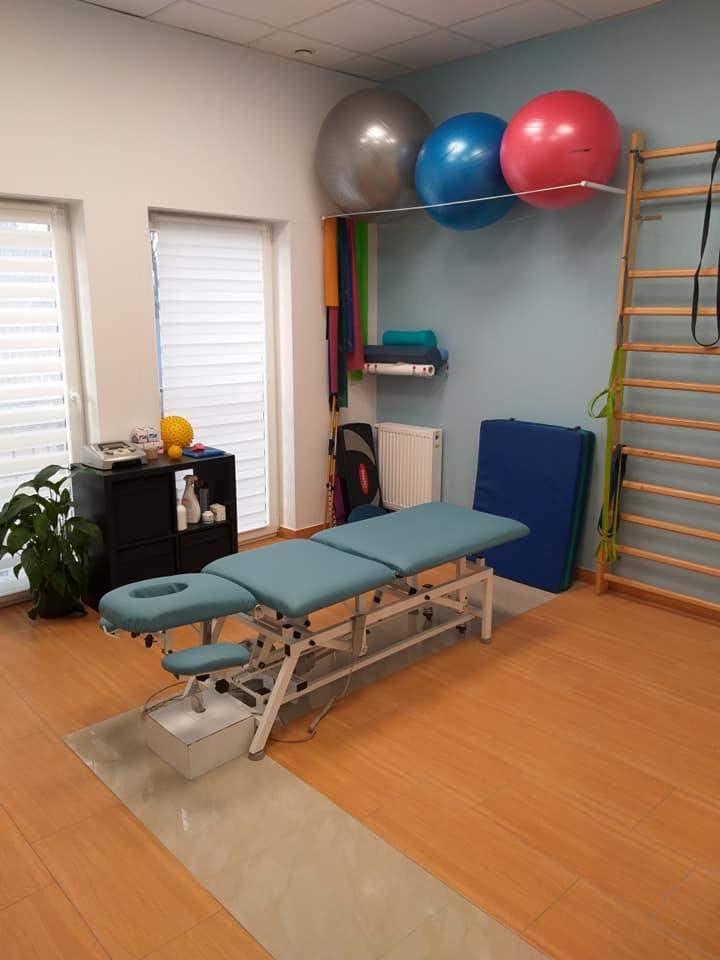 Jasne wnętrze gabinetu z profesjonalnym, regulowanym stołem do zabiegów, drabinką gimnastyczną i kolorowymi piłkami do ćwiczeń na ścianie.