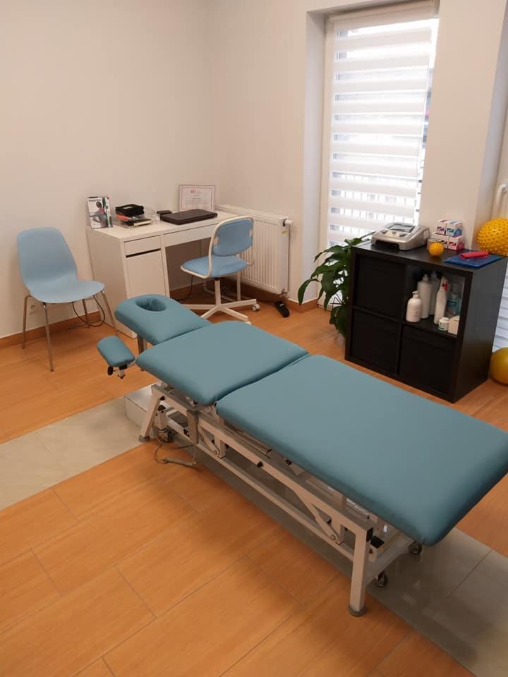 Jasne wnętrze gabinetu z profesjonalnym, regulowanym stołem do zabiegów w kolorze błękitnym, biurkiem z laptopem i certyfikatem na ścianie, oraz szafką z akcesoriami medycznymi.