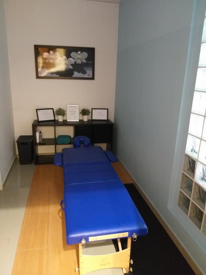 Niebieski stół do zabiegów w jasnym gabinecie z dekoracyjnym obrazem kwiatów i regałem z certyfikatami.