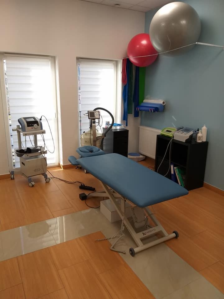 Jasne wnętrze gabinetu z niebieskim łóżkiem do terapii manualnej, sprzętem medycznym na wózkach, piłkami rehabilitacyjnymi na ścianie i drewnianą podłogą.