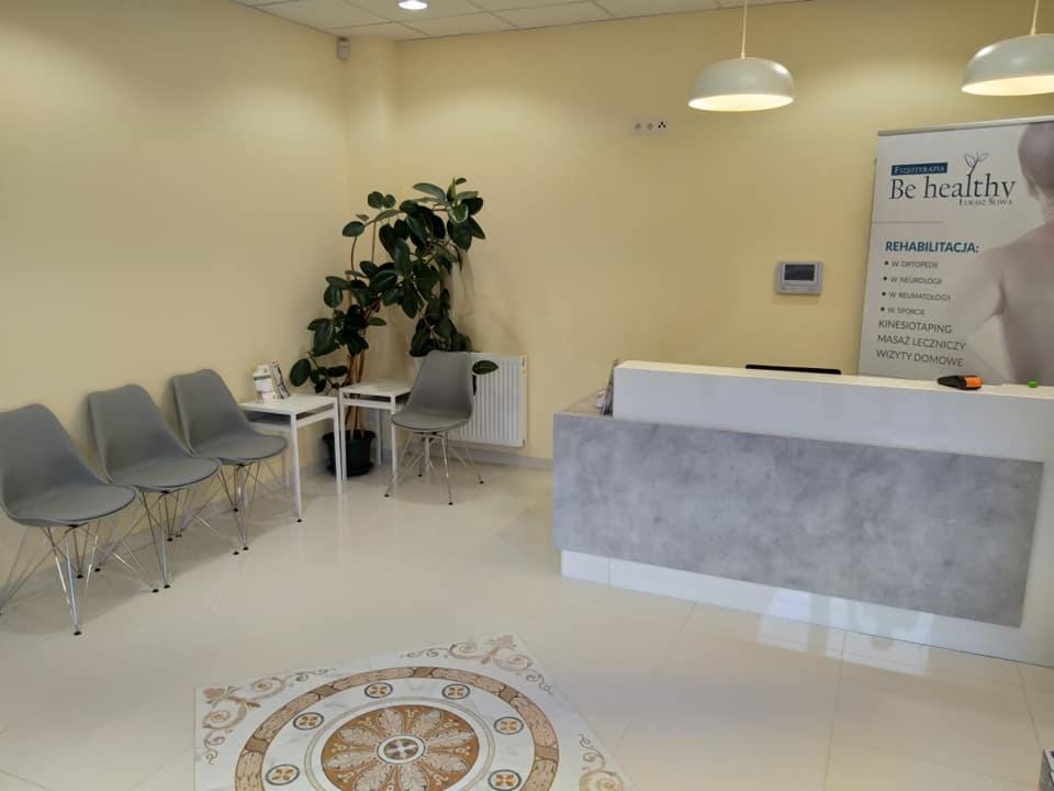 Jasne wnętrze poczekalni gabinetu rehabilitacji z recepcją o betonowej fakturze, czterema szarymi krzesłami, stolikami, rośliną doniczkową i banerem reklamowym z napisem 'Be healthy' i listą usług...