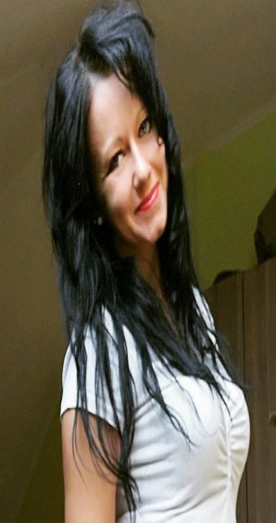 Uśmiechnięta kobieta z długimi, czarnymi włosami w białej bluzce, portret z lekkiego ukosa.