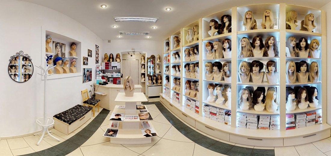 Wnętrze salonu z szerokim wyborem peruk na manekinach, umieszczonych na półkach i w witrynach, z katalogami na stole ekspozycyjnym.