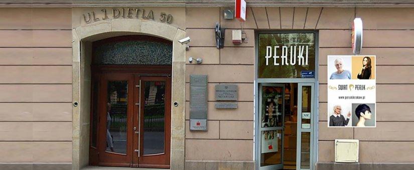 Fasada budynku z dwoma wejściami: jedno z drewnianymi drzwiami pod kamiennym łukiem z adresem 'Ul. I Dietla 50', drugie z napisem 'Peruki' nad szklanymi drzwiami i reklamą peruk po prawej stronie.