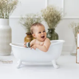 Uśmiechnięty niemowlak w białej wanience, trzymający małą zabawkę, w tle dekoracje z suszonych kwiatów w wazonach i drewniane klocki z literami R, O, K.