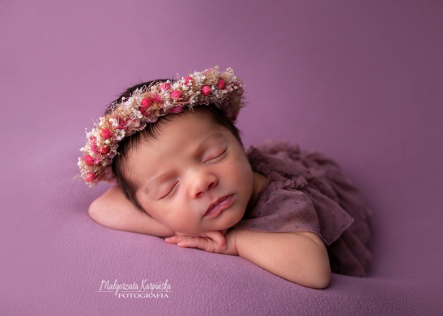 Śpiąca noworodka w fioletowej sukience i wianku z drobnych różowych i białych kwiatów na fioletowym tle.