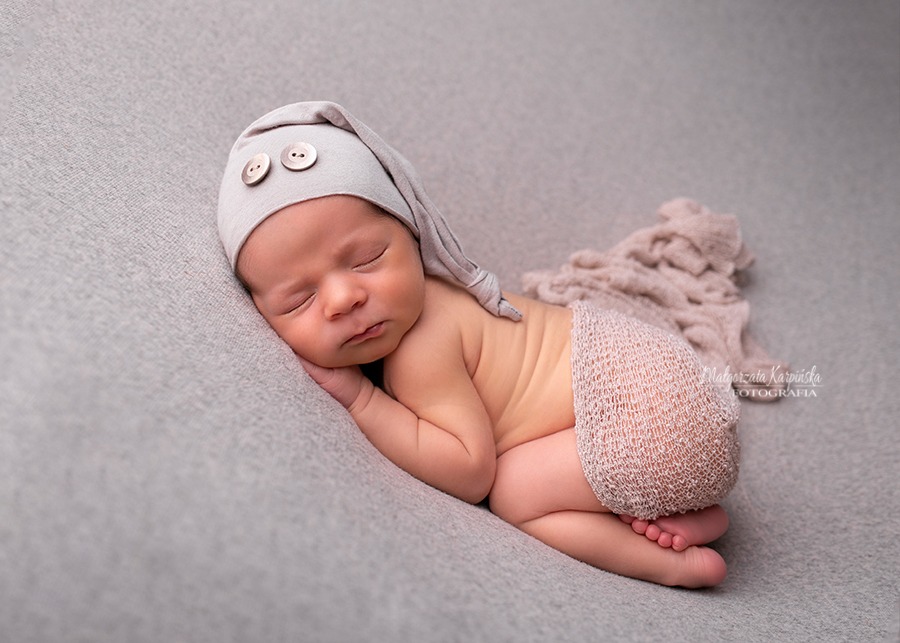 Śpiący noworodek w beżowej czapeczce z dwoma guzikami i ażurowych spodenkach, leżący na szarym tle z delikatnym materiałem obok.